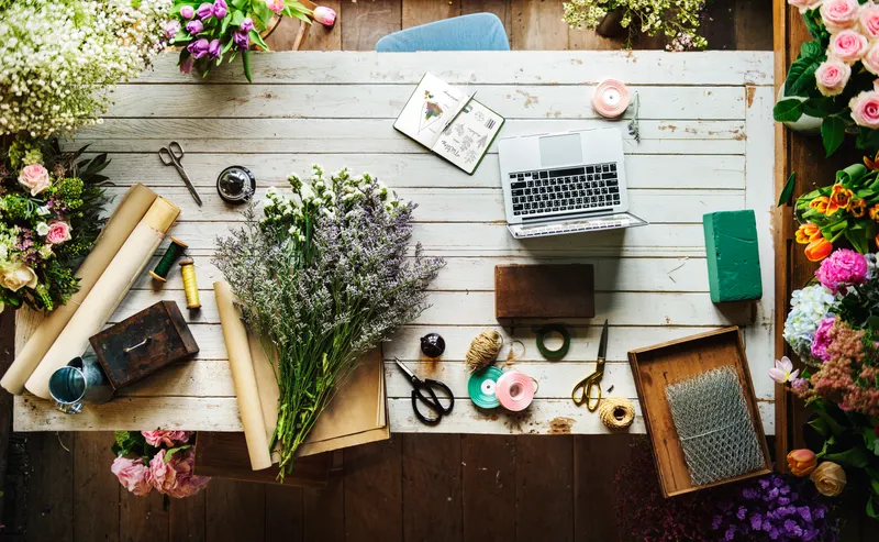 Foto de ejemplo: mesa de florista vista desde arriba con flores sueltas, rollo de papel kraft, cinta y tijeras